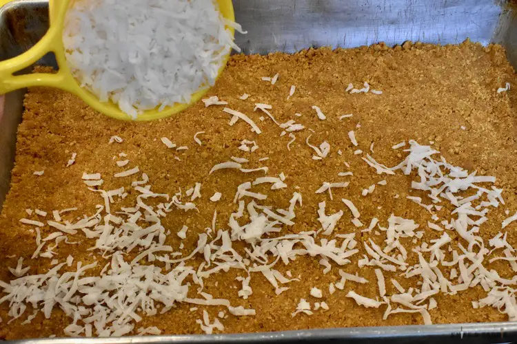Pressed graham crackers crumbs in a 9x13 inch baking pan with sprinkled coconut flakes over the crumbs