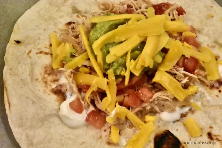 Crockpot chicken tacos in a tortilla with shredded cheddar, salsa, tomatoes, guacamole and sour cream