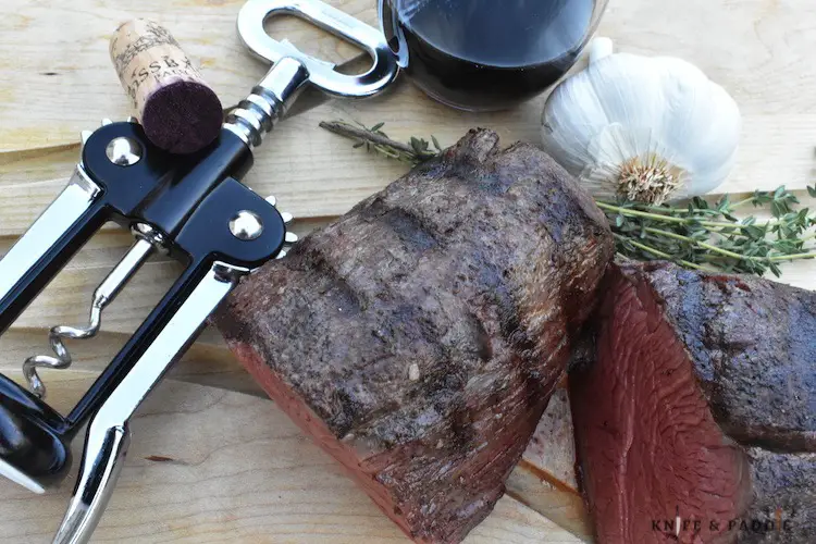 Sous vide beef tenderloin on a cutting board with thyme, garlic, a glass of wine, cork and wine opener
