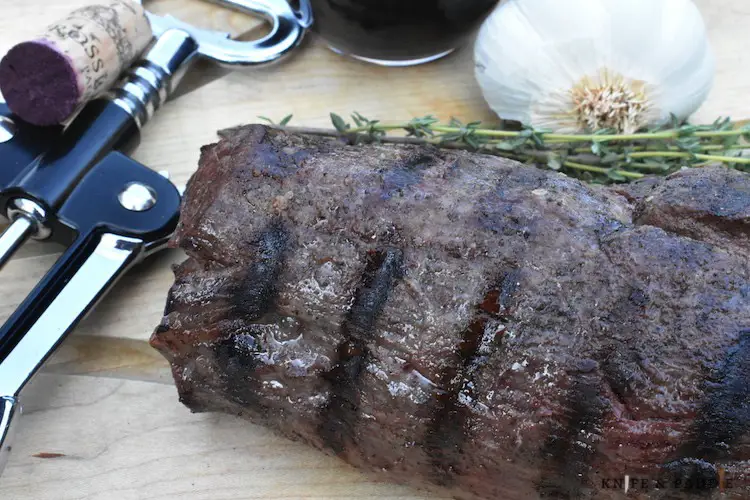 Charred beef tenderloin, thyme, garlic, wine and wine opener on a cutting board