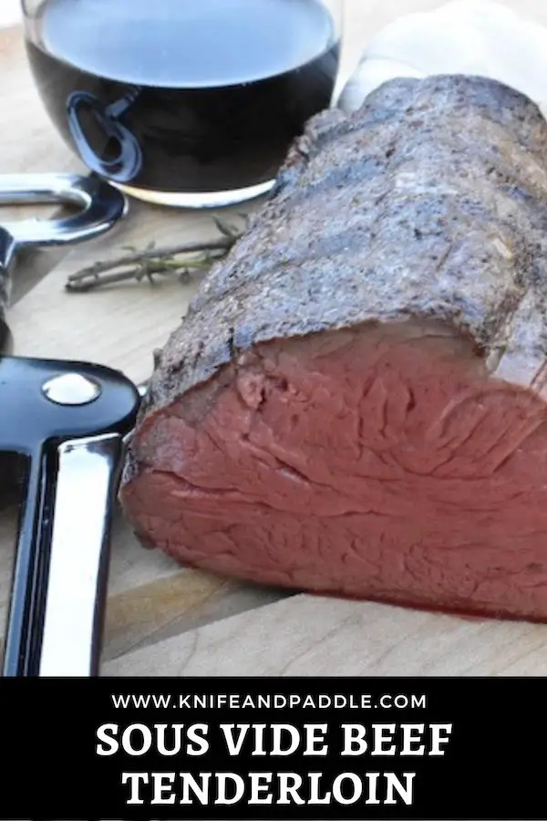 Sous vide beef tenderloin on a cutting board with thyme, garlic a glass of wine, and wine opener