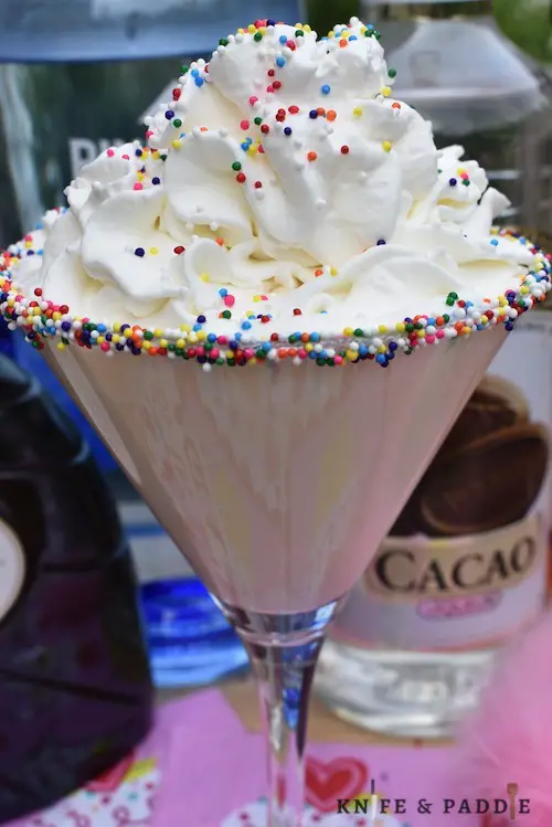Birthday Cake Martini with whipped cream and rainbow nonpareils in a martini glass rimmed with fluff and rainbow nonpareils