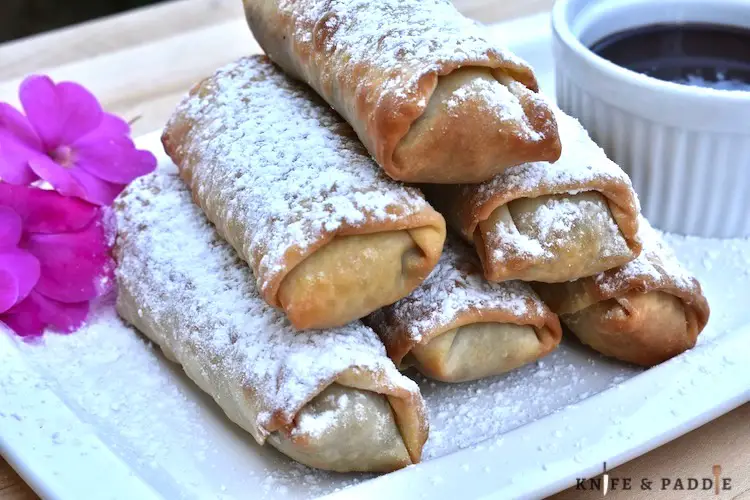 Cookie Dough Egg Rolls stacked on a plate sprinkled with powdered sugar with a side of chocolate dipping sauce