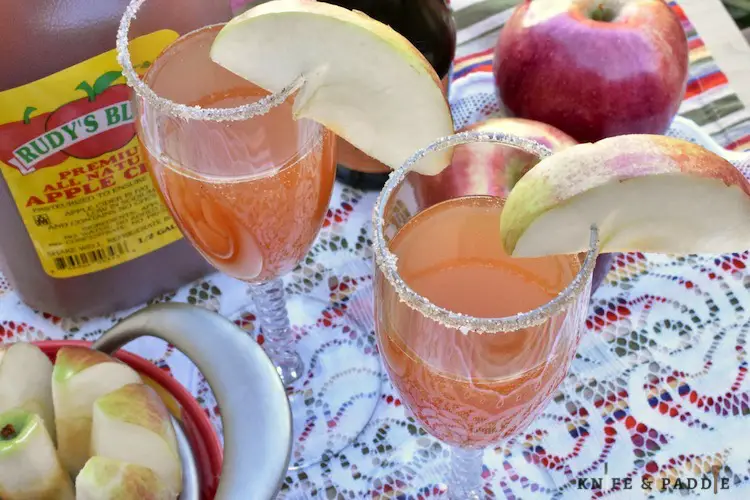 Sliced apples, apple cider, apples and apple cider mimosas in champagne glasses garnished with maple syrup and cinnamon sugar topping and an apple slice