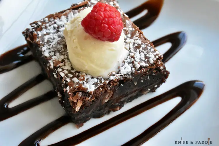 Decadent Fudge Brownies cut into a square on a chocolate drizzled plate topped with ice cream and a raspberry