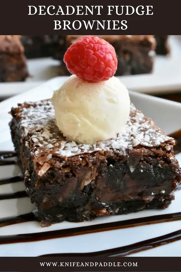 Decadent Fudge Brownies cut into a square on a chocolate drizzled plate topped with ice cream and a raspberry