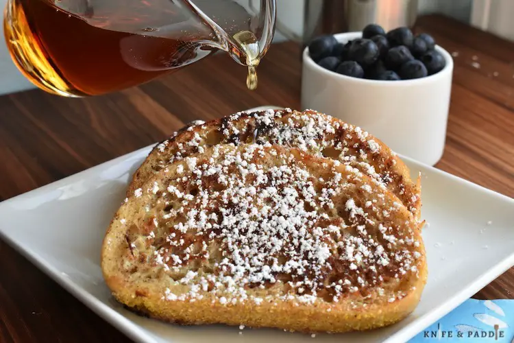 French Toast sprinkled with powdered sugar served on a plate with a side of maple syrup and blueberries