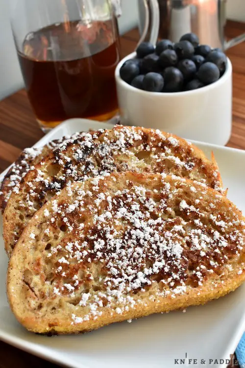 French Toast sprinkled with powdered sugar served on a plate with a side of maple syrup and blueberries