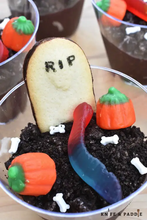 Graveyard treats with chocolate Jello, crushed Oreos, candy pumpkins, candy bones, gummy worms and a RIP tombstone Milano cookie