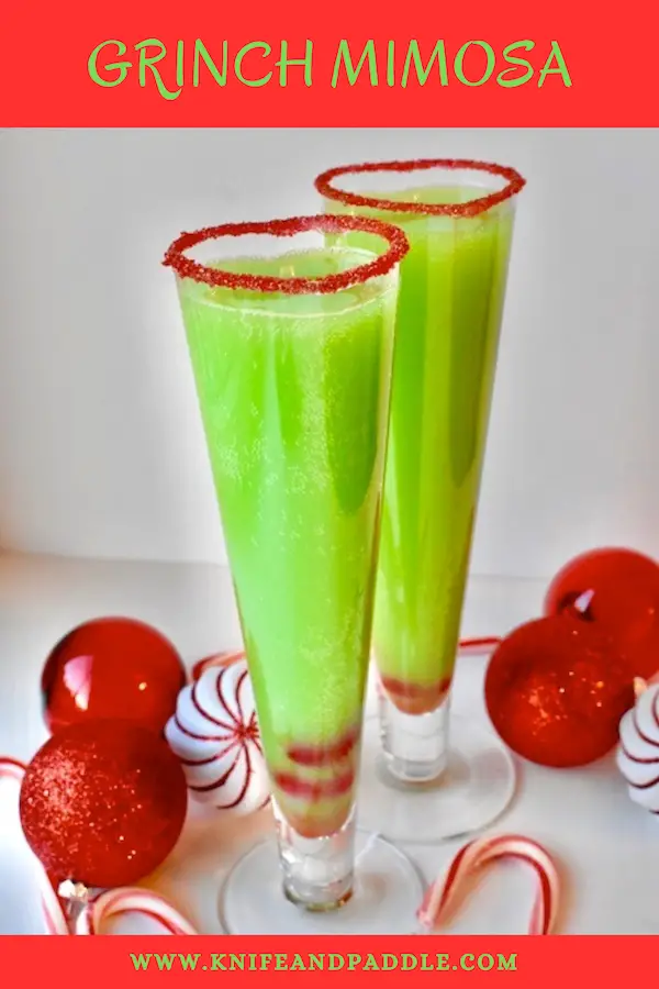 Bright green Grinch Mimosa in a heart shaped champagne glass rimmed with red sparkling sugar and two maraschino cherries in the bottom of the glass