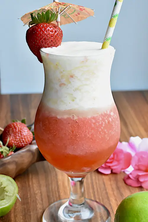 Layered piña colada and strawberry daiquiri cocktail in a hurricane glass garnished with a fresh strawberry and a cocktail umbrella