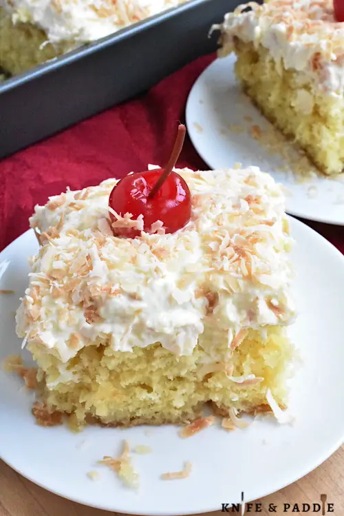 Slice of Pina Colada Poke Cake on a plate topped with whipped topping, toasted coconut and a maraschino cherry