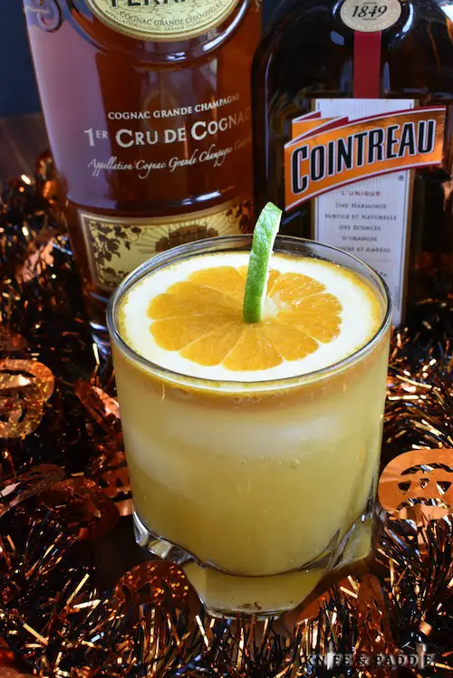 Cognac, Cointreau, orange juice and ginger ale mixed together to create a festive Halloween cocktail garnished with a pumpkin made from a slice of orange and a lime peel