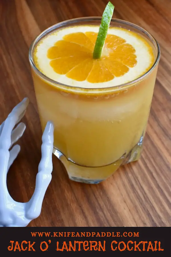 Jack O' Lantern Cocktail made with cognac, cointreau, orange juice and ginger ale mixed together to create a festive Halloween cocktail garnished with a pumpkin made from a slice of orange and a lime peel